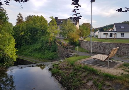 Landschaftsliege aus Holz mit Bach im Vordergrund und dörflicher Bebauung 