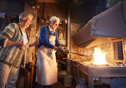 Ein älterer Mann in einer Schürze schmiedet Eisen im Ofen, eine Frau schaut lächelnd zu.