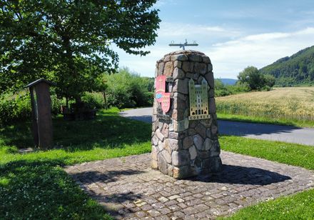 Steinerner Infopunkt am Rand eines Radweges mit angebrachtem Wappen der Ortschaft und Silhouette des Corveyer Westwerkes