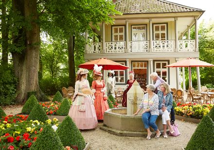 Personen in historischen Kostümen stehen vor einem Café in einem gepflegten Garten mit Blumenbeeten.